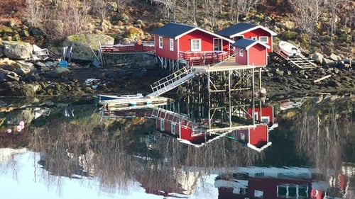 Traditional red cabins stand by the tranquil waters surrounded by snow-capped mountains. Lofoten.