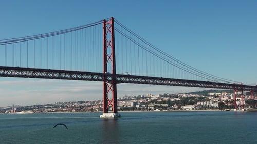 View of a red suspension bridge spanning a river with a vibrant city in the background