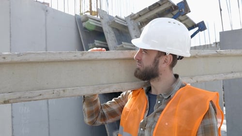 Builder man in hardhat and vest carrying timber on building site in his working day