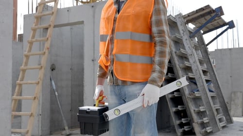 Builder repairman in protective helmet and vest stands at workplace in building and holds ruler