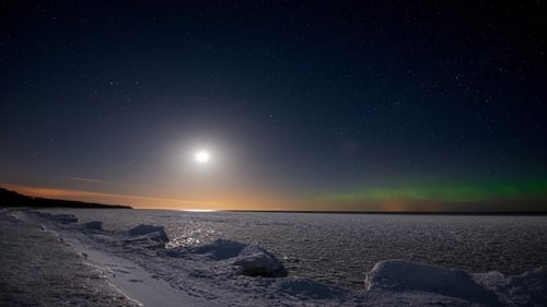 Timelapse. Seashore covered with ice under a starry sky with moon and northern lights.