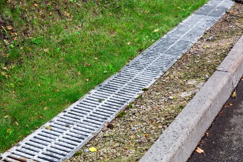 Preview: Drainage System On The Side Of The Road Behind A Gutter With An Iron Grate And Green Grass.