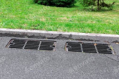 Preview: Two Manhole Cover Rain Grate On An Asphalt Road Near A Stone Curb On The Side Road Green Grass On
