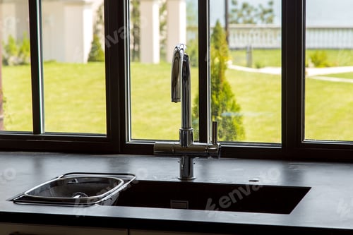 Preview: Chromed Faucet In The Kitchen Sink With A Black Stone Tabletop Near The Window With A View Of The