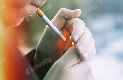 Preview: A Caucasian Man Is Lighting Up A Cigarette Outdoors. Focus Point Is On The Cigarette. Image Has A