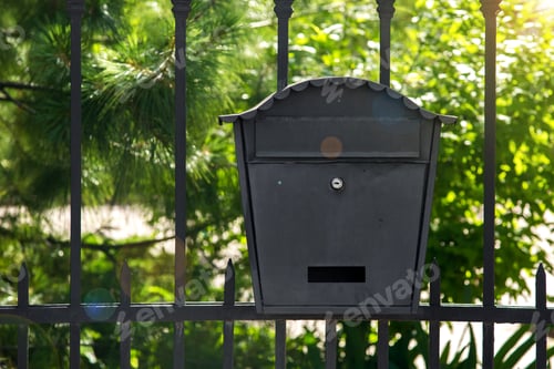 Preview: Black Iron Mailbox Mounted On A Metal Fence On A Green Plan With Green Trees On A Sunny Day.