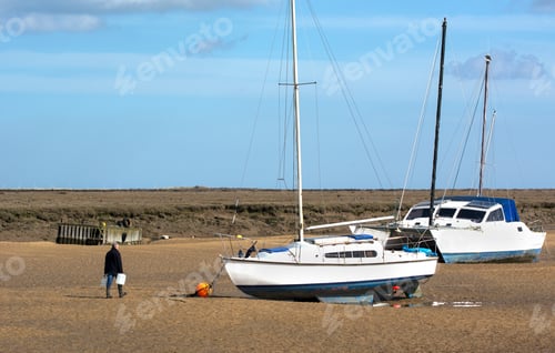 Preview: Solitary Man Walking Towards Small Boats Stranded At Low Tide At Wells Next The Sea, Norfolk, Uk