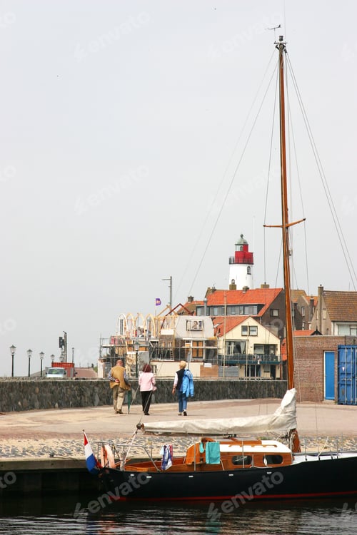 Preview: Sailing Ship Anchored In Harbor In Front Of Fishing Village Lighthouse, People Walking To Town