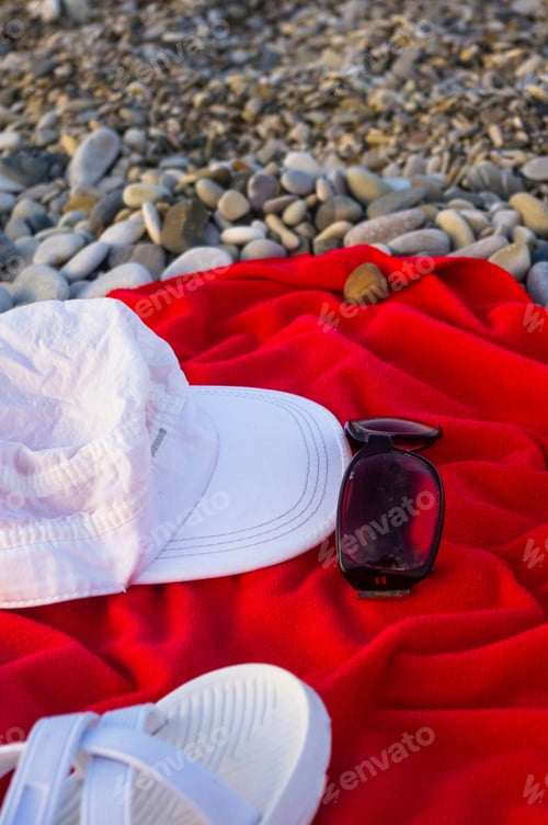 Preview: Red Towel, White Cap, Sunglasses And Beach Slippers On Pebble Beach, Sunset
