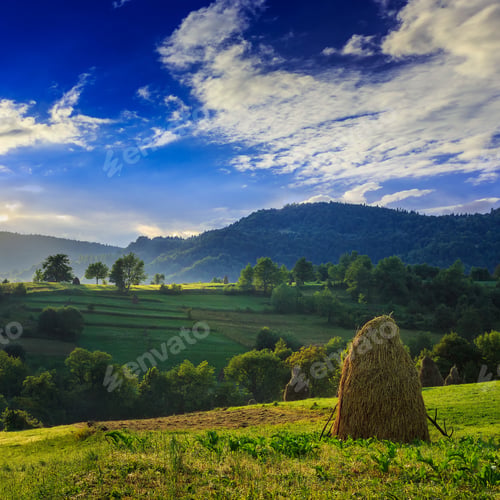 Preview: Stack Of Hay On A Green Meadow In The Mountains In The Morning Under A Blue Summer Sky