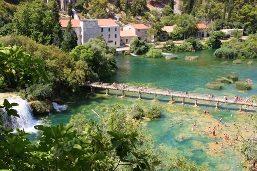 Preview: People Swimming In Natural Swimming Pool With Clear Water, Waterfalls, Bridge And Village