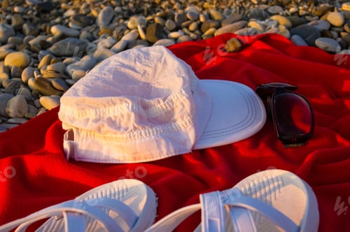 Preview: Red Towel, White Cap, Sunglasses And Beach Slippers On Pebble Beach, Sunset