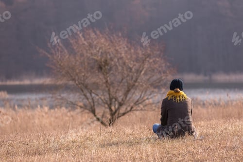 Preview: Young Woman Sitting On Grass On Meadow Near Lake And Dreaming. Photo Taken From Behind.
