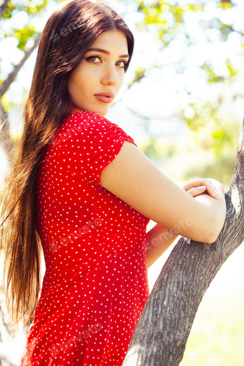 Preview: Pretty Young Woman In Red Dress Smiling Cheerful In Green Park At Tree On Summer Sunny Day