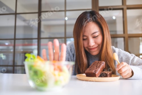 Preview: A Woman Choosing To Eat Brownie Cake And Making Hand Sign To Refuse A Vegetables Salad On The Table