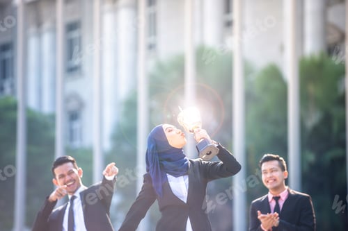 Preview: Successful Muslim Business Woman Kissing A Trophy And Celebrating, With Male Teammates Supporting