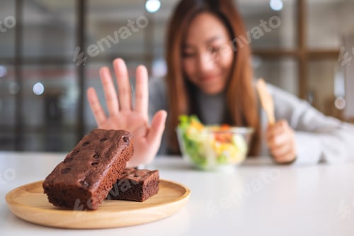 Preview: A Woman Choosing To Eat Vegetables Salad And Making Hand Sign To Refuse A Brownie Cake On The Table
