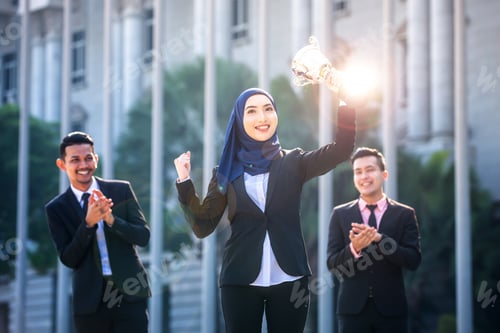 Preview: Successful Muslim Business Woman With Arms Up Holding A Trophy And Celebrating, With Teammates