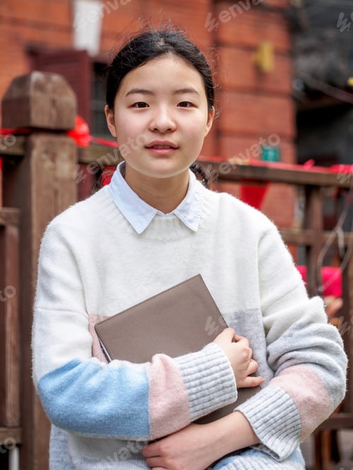 Preview: Portrait Of A Young Beautiful Chinese Girl Hold A Notebook And Smile At Camera, Against Blur School