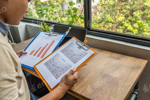 Preview: Asian Freelancer Holding And Analyzing Statistic Documents At Rustic Workplace