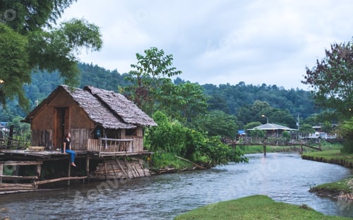 Preview: Portrait Image Of A Woman Sitting On Cabin House Balcony By The River With Mountains And Nature