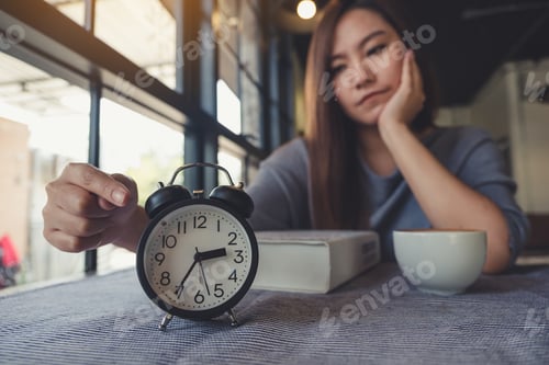 Preview: A Beautiful Asian Woman Pointing At A Black Alarm Clock On Table With Feeling Bored
