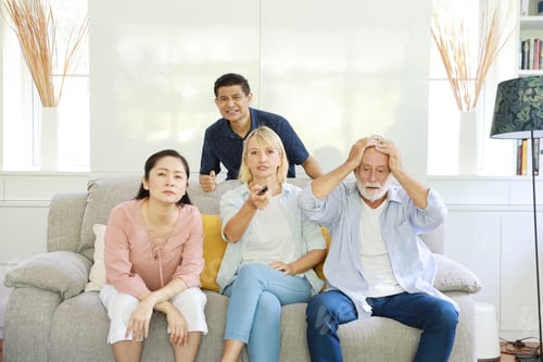 Visualização: Família multiétnica de idosos infelizes sentados e assistindo televisão no sofá cinza na sala de estar.
Eles