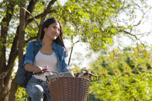Preview: Asian Female Student Riding A Bicycle In A Park On The Campus Filled With Trees.
