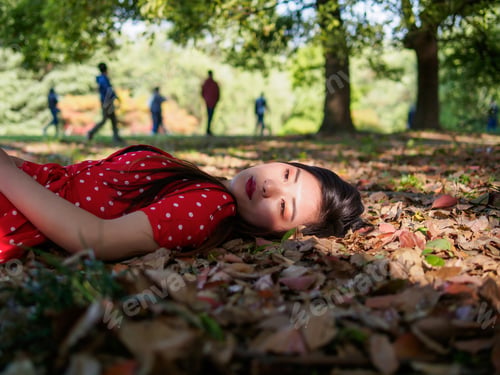 Preview: Beautiful Young Woman In Red Dress Lying On Ground In Forest Park With Blur People Walking