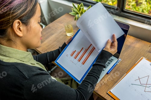 Preview: Female Asian Freelancer Reading Diagram Business Document At Rustic Workplace