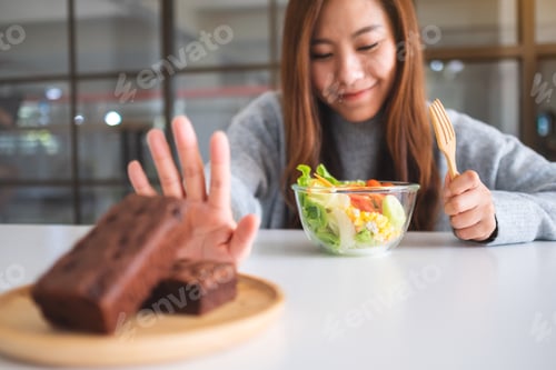 Preview: A Woman Choosing To Eat Vegetables Salad And Making Hand Sign To Refuse A Brownie Cake On The Table
