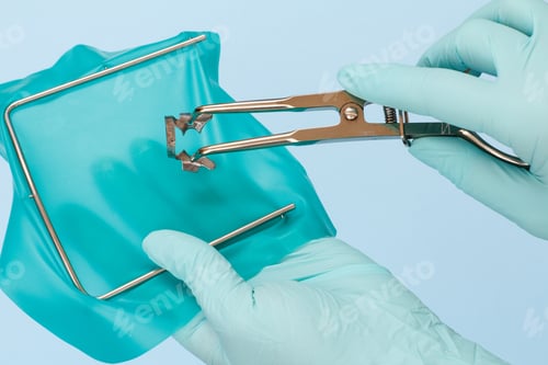 Preview: Dentist'S Hands In Latex Gloves With Dental Tongs, Cofferdam Scarf And Metal Frame On Blue