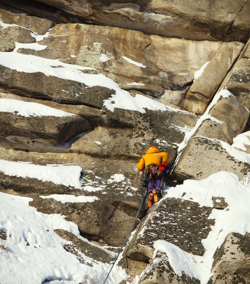 Preview: Mountaineer Figure In Bright Clothes On A Sunlit Snow-Covered Rock Wall During A Winter Ascent In