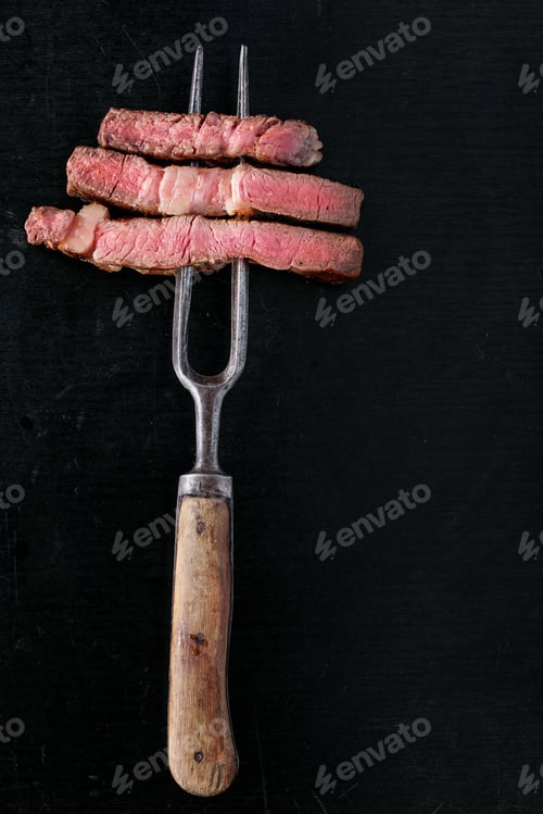 Preview: Grilled Striploin Angus Steak Cut In Pieces On The Fork On Black Wooden Board. Top View. Copy Space