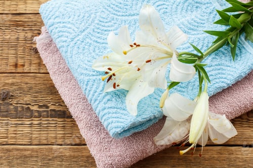 Preview: Blue And Pink Soft Terry Towels With Bouquet Of White Lily Flowers On Old Wooden Boards. Top View.