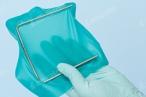 Preview: Dentist'S Hand In Latex Glove With Dental Cofferdam Scarf And Metal Frame On Blue Background