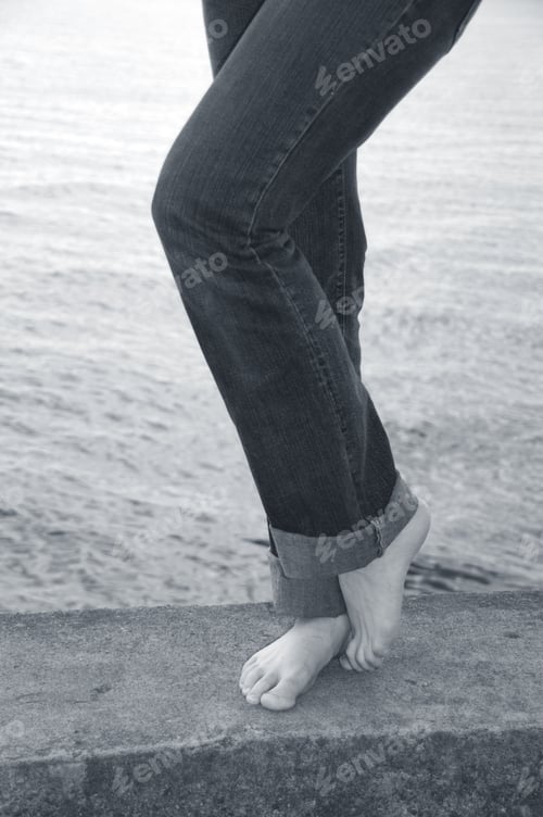 Preview: A Woman'S Legs In Jeans With Bare Feet Standing On A Cement Pier At The Beach.