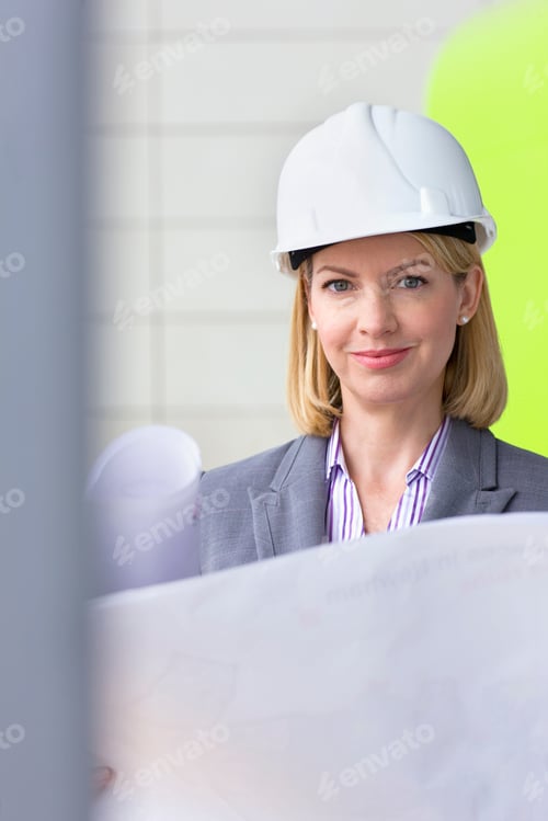 Preview: Female Architect In Hard Hat Looking At Blueprint In Modern Building Looking At Camera