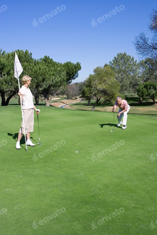Aperçu: Mature Couple Playing Golf, Man Lining Up Golf Shot On Putting Green, Woman Holding Flag