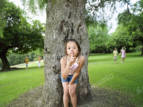 Preview: Girl With Finger On Lips Playing Hide And Seek In Park, Hiding From Friends Behind Tree