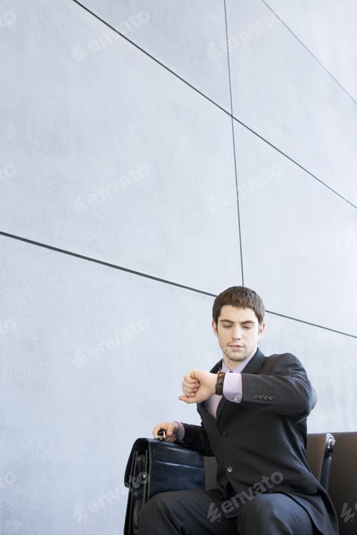 Preview: A Businessman Sitting Down Looking At His Watch Holding A Briefcase