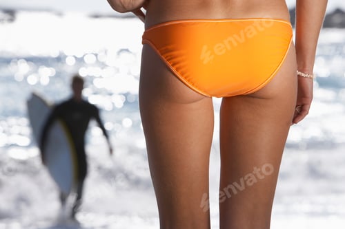 Preview: Young Woman In Orange Bikini Watching Surfer In Sea, Back View