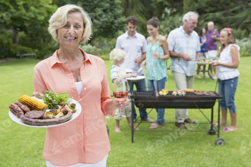 Preview: Portrait Of Smiling Senior Woman Holding Plate Of Barbecue And Wine With Family In Background