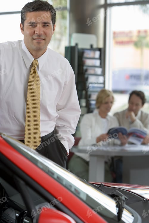 Preview: Couple Looking At Brochure In Car Showroom, Focus On Salesman In Foreground, Smiling, Portrait