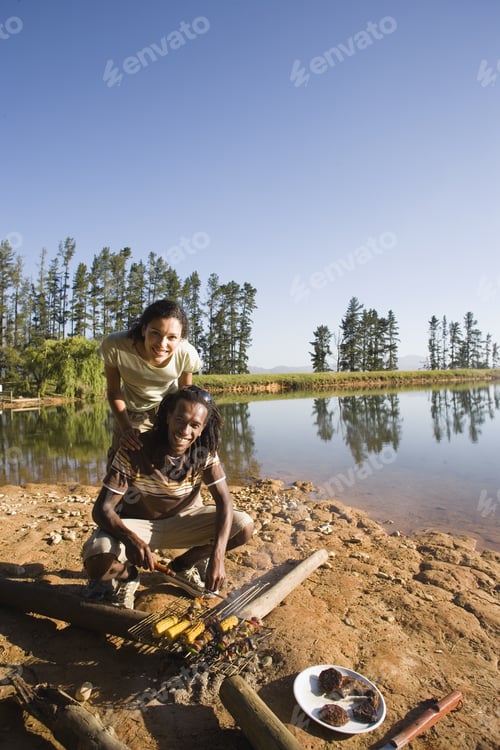 Preview: Couple Cooking Food On Lakeside Camping Trip, Man Grilling Corncobs On Campfire, Smiling, Portrait