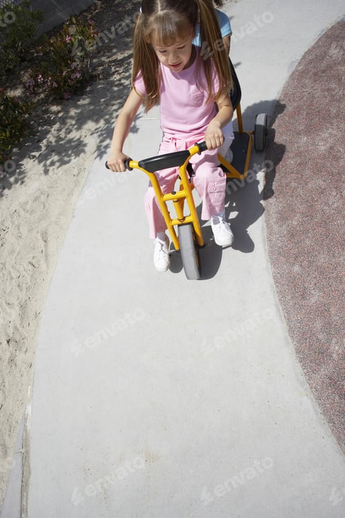 Preview: Two Girls (4-6) Riding Toy Tricycle In Playground, Front View, Elevated View (Tilt)