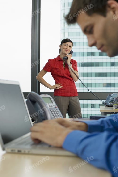 Preview: Businessman Using Laptop In Office, Focus On Woman Using Telephone In Background, Smiling, Side View