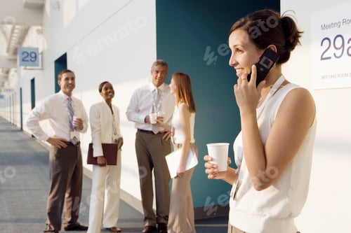 Preview: Four Business People Talking In Corridor Outside Meeting Room, Focus On Businesswoman Using Mobile