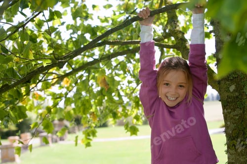 Preview: Portrait Of Smiling Young Girl Playing In Garden Climbing Tree In Countryside Hanging From Branch