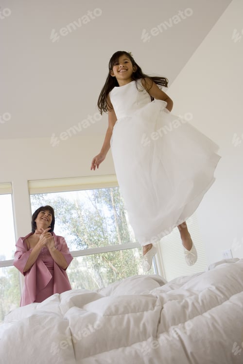 Preview: Senior Woman Watching Girl (8-10), Dressed In Bridesmaid'S Dress, Jumping On Bed In Bedroom